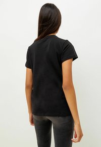 Kvinna med långt mörkt hår som bär en enkel svart kortärmad T-shirt och mörkgrå jeans, stående vänd bort från kameran mot en vit bakgrund.