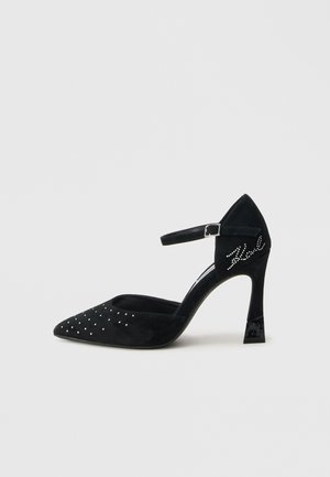 Μαύρο σουέτ ψηλοτάκουνο παπούτσι με μυτερή μύτη, λουράκι στον αστράγαλο και διακοσμητικά καρφιά στο μπροστινό μέρος· υφασμένος τακούνι με γυαλιστερή λεπτομέρεια.