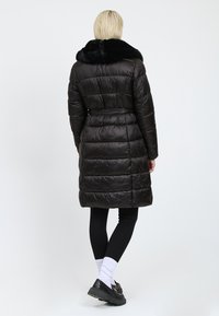 Cappotto puffer nero con colletto in pelliccia artificiale, vita con cintura e texture trapuntata, che arriva a metà coscia, con maniche lunghe e tasche laterali.