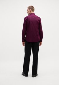 Langermet, burgunder skjorte med spiss krage og knappmansjetter, brukt med svarte bukser. Har et glatt, litt teksturert stoff.