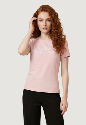 Jeune femme aux longs cheveux roux bouclés portant un t-shirt rose clair GUESS et un pantalon noir, posant avec une main touchant ses cheveux.