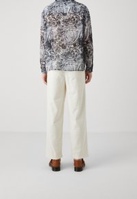 Camicia leggera e semi-trasparente a maniche lunghe con un motivo astratto in nero, grigio e bianco, abbinata a pantaloni color crema e calzature marroni.