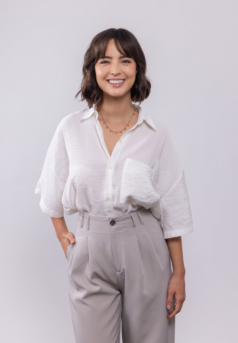 Witte, gestructureerde overhemd met knopen en een borstzak, gecombineerd met hooggesneden, lichtgrijze geplooide broek. Eenvoudige gouden ketting als accent.
