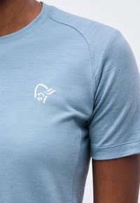 Φωτεινό γαλάζιο κοντομάνικο μπλουζάκι με στρογγυλή λαιμόκοψη σε άτομο, με μικρό λευκό μινιμαλιστικό λογότυπο προσώπου στο αριστερό στήθος.