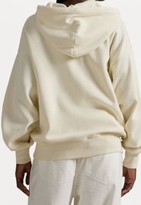 Kremowa, oversizowa bluza z kapturem ze sznurkiem, ściągaczami na rękawach i miękką, szczotkowaną fakturą, zestawiona z jasnokremowymi spodniami z tylnymi kieszeniami.