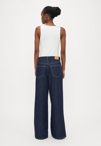 Platām kājām tumši zilas džinsa bikses ar aizmugurējām kabatām, kas pie vidukļa izrotātas ar brūnas ādas logotipa uzlāpi un gludu, fakturētu apdari.