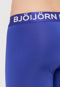 Niebieskie slipy w formie bokserów z gładkiego, elastycznego materiału oraz szerokim, kontrastowym białym paskiem z logo, zaprojektowane z myślą o wygodzie i dopasowaniu.