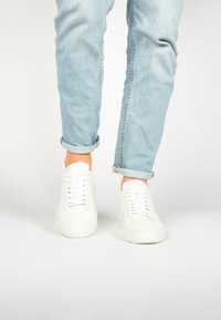 Baskets en cuir blanc avec une texture lisse, dotées d'un système de laçage et de bouts arrondis, portées avec un jean bleu clair retroussé.