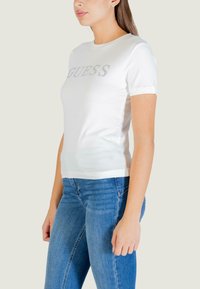 Maglietta bianca a maniche corte con logo "GUESS" in strass, design aderente, abbinata a jeans blu. Materiale in cotone liscio.