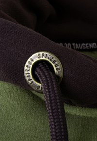 Brown fabric with a smooth texture, featuring a metal grommet and dark braided cord. Underneath, a green fabric shows a soft texture.