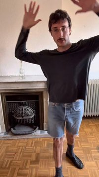 Szemüveges, bajszos férfi fekete hosszú ujjú ingben és farmer rövidnadrágban, kezeit felemelve áll egy kandallós, fa padlós szobában.