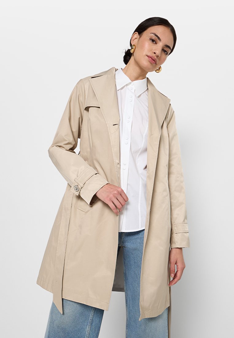 Manteau beige long en tissu lisse, présentant un grand col, des poignets boutonnés et des poches latérales, porté sur une chemise blanche et un jean.