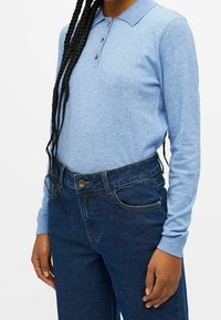 Personne portant un polo à manches longues bleu clair, rentré dans un jean taille haute bleu foncé, avec des cheveux tressés tombant sur l'épaule.