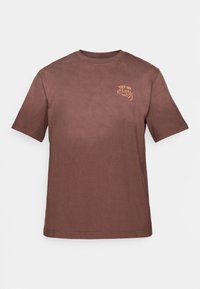 Καφέ κοντομάνικο βαμβακερό T-shirt με μικρή πορτοκαλί γραφική παράσταση της λέξης "Karma" και δύο σχήματα φωτοστέφανου στο αριστερό στήθος.