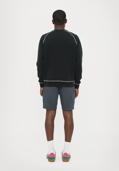 Svart sweatshirt med raglanärmar, grå sömmar; kombinerad med mörkblå shorts. Modellen bär vita strumpor och flerfärgade sneakers.