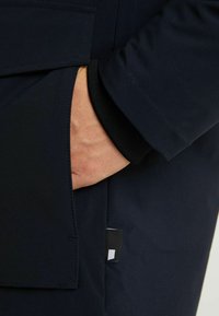 Granatowa odzież wierzchnia z ręką w bocznej kieszeni, detalem z prążkowanym mankietem i przeszyciami. Mała metka z logo widoczna obok kieszeni.