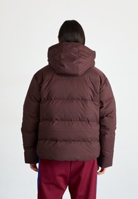 Smeđi puffa jakna s horizontalnim quilting dizajnom i kapuljačom. Ima gustu, podstavljenu teksturu. Stražnji pogled pokazuje elastičnu obrubu odjeće.
