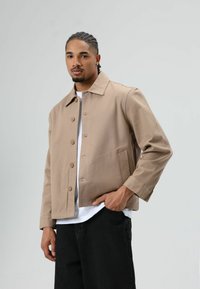 Beige, knoopjasje met een spreidekraag, borstzakken en lange mouwen. Draagt over een wit T-shirt en gecombineerd met zwarte broek.