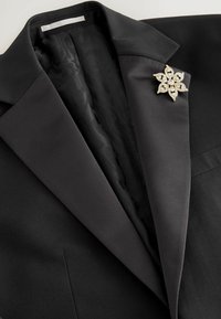 Schwarzer Blazer mit Satinrevers und einer goldenen und kristallklaren Blumenbrosche auf der linken Seite.