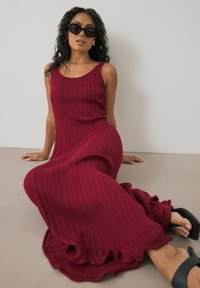 Bordeauxfarbenes, geripptes Maxikleid mit figurnahem Oberteil und ausgestelltem Rüschensaum. Getragen mit schwarzen Sonnenbrillen und Rutschsandalen. Glatte Textur.