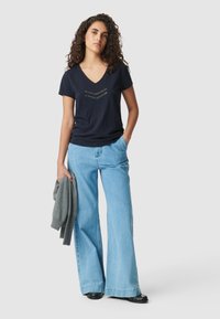 Mujer joven con cabello rizado, vistiendo una camiseta de cuello en V azul marino con "1987", pantalones vaqueros de pierna ancha azul claro, zapatos negros, sosteniendo un suéter gris.