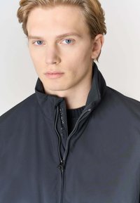 Giacca nera con zip e collo alto, realizzata in tessuto liscio e opaco; presenta una chiusura con zip e un design interno a maglione.