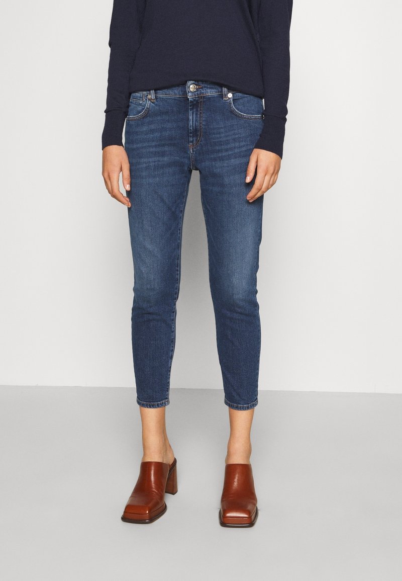 Femme portant un jean court bleu, des mocassins marron à talon et un haut sombre à manches longues, debout devant un fond clair uni.