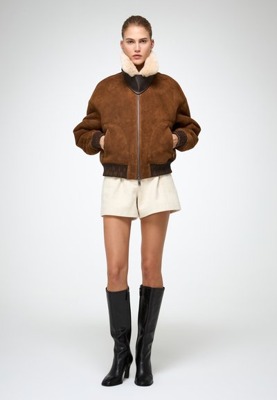 Giacca bomber in suede marrone con colletto scuro, dotata di polsini a costine, tasche frontali e zip. Indossata sopra pantaloni corti bianchi e stivali neri al ginocchio.