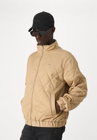 Beige tikandatud jakk kõrge kaelusega, elastsete mansettide ja küljetaskutega. Materjal näeb välja pehme ja sileda tekstuuriga.