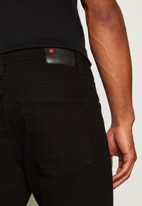 Svarta denimjeans med en slät textur, prydda med en läderetikett prentad med "DENIM PROJECT" och en röd prick. Passformen är snug runt midjan.