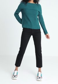 Pull en tricot vert sarcelle avec design épaules dénudées, associé à un jean noir court et des baskets multicolores. Tissu texturé et coupe droite.