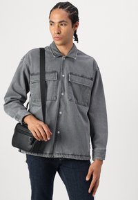 Giacca di denim grigia con due tasche sul petto e bottoni a pressione argentati. Il modello tiene una borsa a tracolla nera. Jeans scuri completano il look.