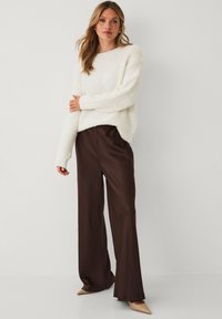 Kvinde står iført en hvid fuzzy sweater, mørkebrune vide bukser og beige spidse hæle mod en ensfarvet baggrund.