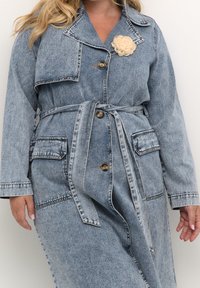 Žena v světle modré džínové bundě s páskem, velkými knoflíky, kapsami na klopách a krémovou květinovou broží na hrudi.