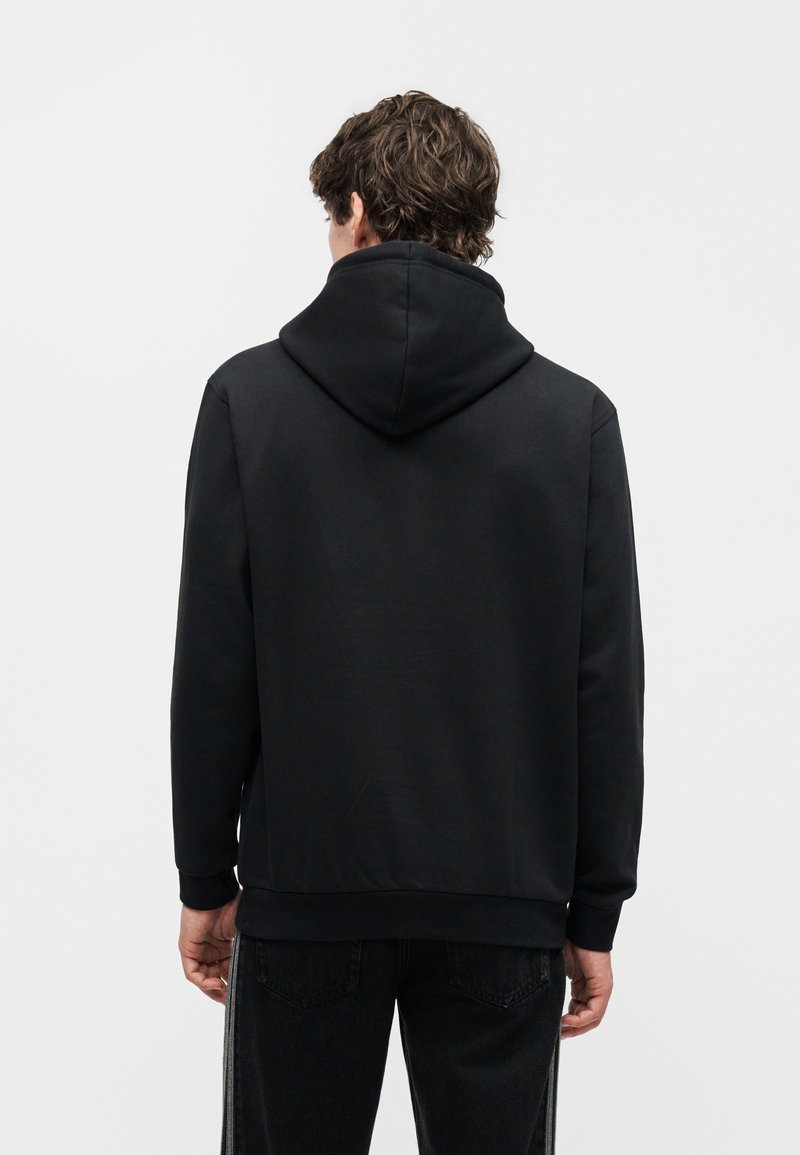 Mann mit lockigem Haar, der einen schlichten schwarzen Hoodie und schwarze Hose trägt, steht mit dem Rücken zu einem weißen Hintergrund.