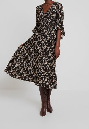 Frau trägt ein schwarzes Midikleid mit beigem Blumenmuster und dreiviertellangen, gerafften Ärmeln, kombiniert mit dunkelbraunen kniehohen Stiefeln.