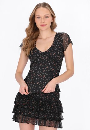 Ung kvinde med bølget hår iført en sort blomsterprint kjole med korte ærmer og lagdelt, flæset nederdel, stående foran en hvid baggrund.