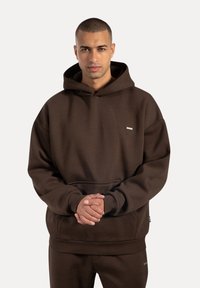 Man som bär en mörkbrun hoodie och matchande byxor, står med händerna sammanfognade framför sig, mot en enfärgad ljus bakgrund.