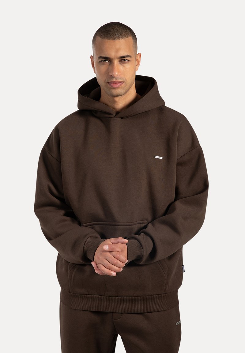 Man som bär en mörkbrun hoodie och matchande byxor, står med händerna sammanfognade framför sig, mot en enfärgad ljus bakgrund.