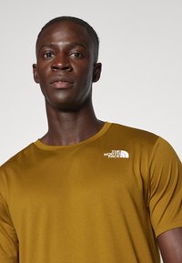 T-shirt de manga curta, com decote redondo, em tecido amarelo mostarda. Apresenta um logotipo branco no lado esquerdo do peito. Textura suave, design ajustado.