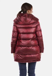 Piumino rosso con cappuccio, caratterizzato da una texture lucida e un design trapuntato orizzontale. Le maniche sono lunghe e ha una lunghezza media.