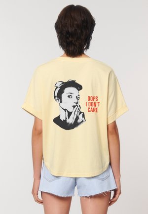 Light yellow t-shirt features a black and white graphic of a woman with a finger to her lips and the text "OOPS I DON'T CARE" in orange.