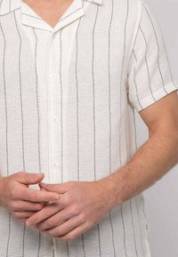 Chemise blanche à manches courtes, avec des rayures verticales noires, fabriquée en tissu texturé, dotée d'un col et d'une fermeture à boutons sur le devant.