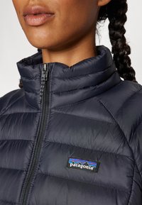 Marineblauwe pufferjack met een hoge kraag, voorzien van lichte gewatteerde secties en een Patagonia logo label op de borst. Zichtbare ritssluiting.