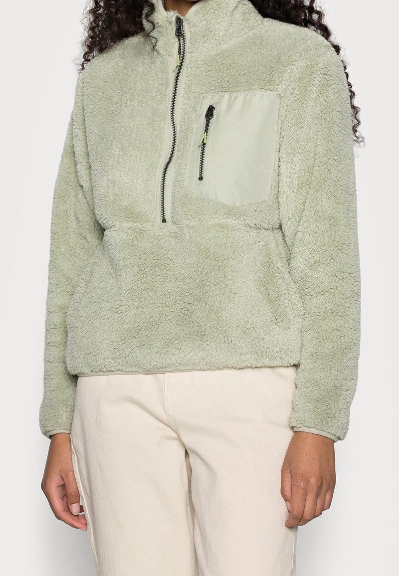 Pull polaire vert clair avec demi-fermeture éclair et poche poitrine zippée, porté avec un pantalon beige clair, montrant le haut du torse et les bras.