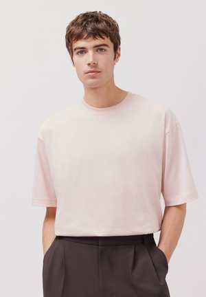 T-shirt rose clair à manches courtes avec un col rond, fabriqué à partir d'un tissu doux ; porté avec un pantalon sombre, affichant une coupe décontractée.