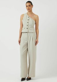 Beige, op maat gemaakte, één-schouder top met knopen en zakken, gecombineerd met ruime, geplooide broeken. Gelaagd stof, strakke design.