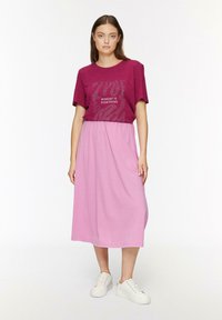 Jupe midi rose à coupe décontractée, associée à un t-shirt bordeaux à manches courtes comportant le texte "L'état d'esprit est tout" dans un design ludique.