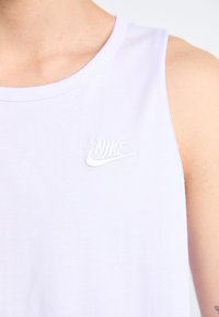 Balta be rankovių maikutė su apvaliu iškirpimu, kuri turi siuvinėtą Nike logotipą kairėje pusėje. Lygus, lengvas audinio tekstūra.
