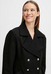 Manteau en laine noir mélangé avec un devant croisé, de grands revers à cran et des boutons ronds brillants. Design classique et structuré.